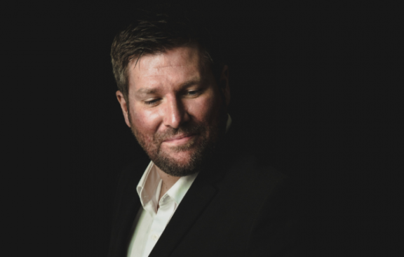 Picture of a man in a black suit and a white shirt with a black background.