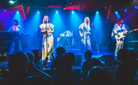 Two women singing, a man plays guitar and another man plays the keyboard on stage.