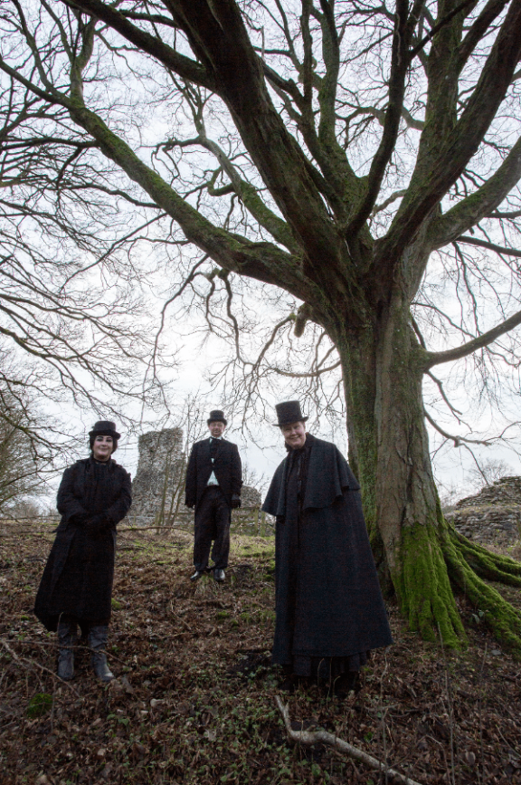 1 lady and 2 men dressed in old fashioned black clothing stand next to a tree