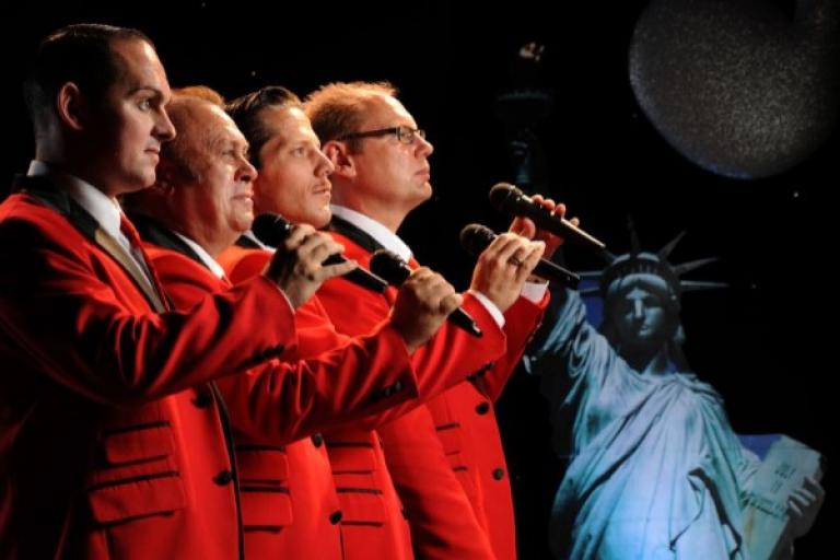 Four men in red jackets singing with a picture of the Statue of Liberty in the background.